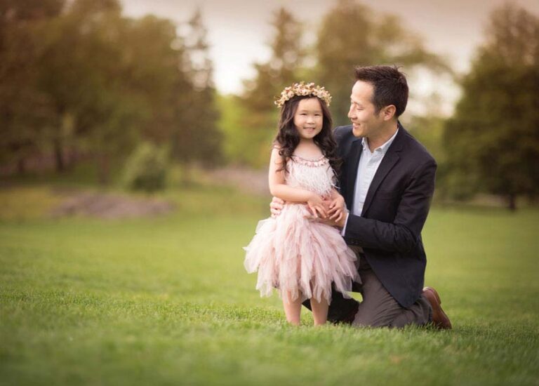 A father kneels on the grass, smiling at his young daughter standing beside him. The girl is wearing a pink, frilly dress and a floral headband. They are in a park with green trees and a soft-focus background. The scene is warm and joyful.
