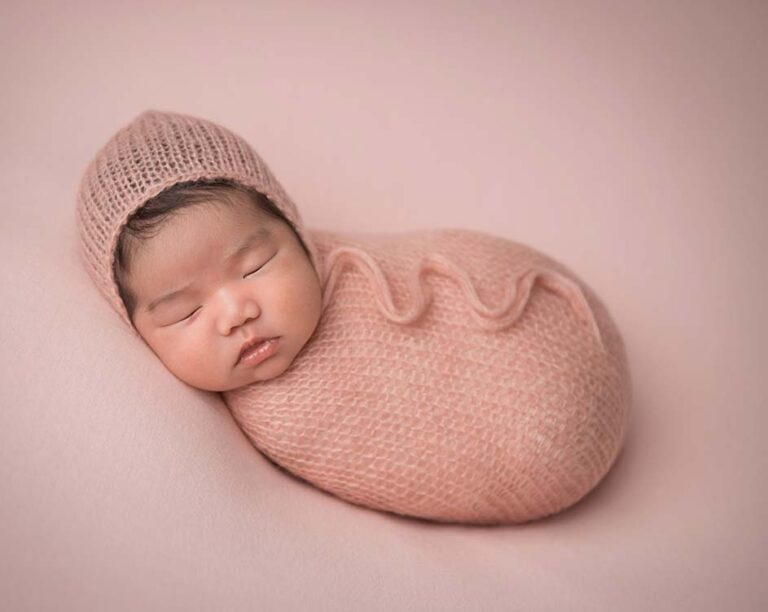 A newborn infant is peacefully sleeping while swaddled in a blush pink knitted blanket with a matching knitted hat. The baby is lying on a soft pink surface, creating a serene and cozy atmosphere.