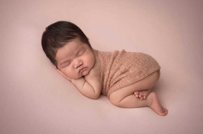 A newborn baby, wrapped in a light brown knitted wrap, sleeps peacefully on a soft pink blanket. The baby is lying on their side with their head resting on one hand and legs slightly curled up. The background is a soft pink color, creating a calming ambiance.