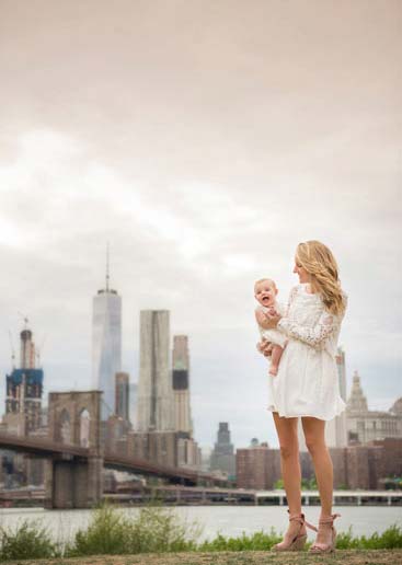A woman in a white dress stands near a waterfront, holding a smiling baby. The skyline of a large city with tall buildings, including the Brooklyn Bridge, is visible in the background. The sky is overcast, creating a serene atmosphere.