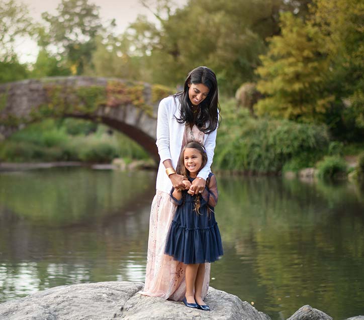 A mother and her young daughter stand on a large rock by a serene lake in Central Park. The woman, dressed in a light, long dress and cardigan, holds the girl's hands from behind. The girl, wearing a navy dress, smiles brightly. A stone bridge and trees are visible in the background.