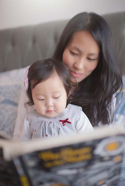 A mother with long dark hair is sitting on a bed, holding and reading a book with her baby daughter. The baby girl, adorned with a small pink bow in her hair and wearing a grey dress, is focused on the book. They share a tender, quiet moment together.