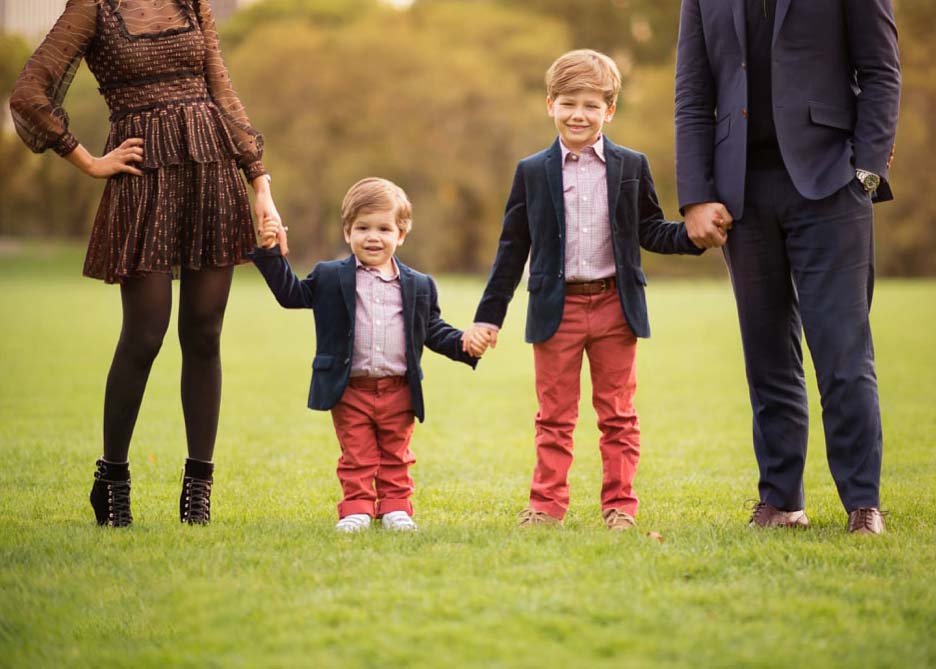 A family of four stands on a grassy field in Central Park. Two siblings are in the middle, holding hands, both wearing blazers and red pants. The parents, partially visible, are holding the children’s hands. Everyone is dressed in semi-formal attire. Trees are in the background.