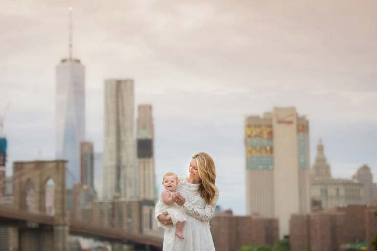 A woman in a white dress cradles a baby in her arms while standing outdoors. The backdrop features several tall buildings and the Brooklyn Bridge, unmistakably placing them in NYC. The sky is cloudy, giving the scene a soft, pastel ambiance.