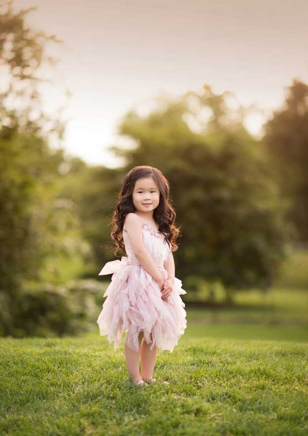 A young girl with long dark hair stands on a grassy field in an NYC park, wearing a fluffy, light pink dress. She smiles softly while holding her hands together in front of her. The background is blurred with greenery and warm, golden light.