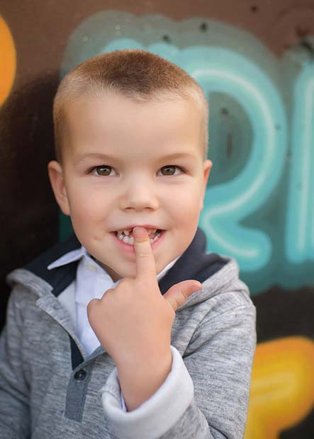 A young boy with short hair smiles and points to his front teeth with his index finger. He is wearing a gray hoodie over a white shirt and stands in front of a colorful graffiti wall.