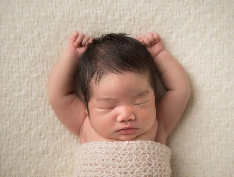 A sleeping baby with dark hair is lying on a cream-colored blanket. The baby's arms are stretched upward, resting on the blanket. The baby is wrapped in a light-colored knit swaddle. The expression on the baby's face is peaceful and serene.
