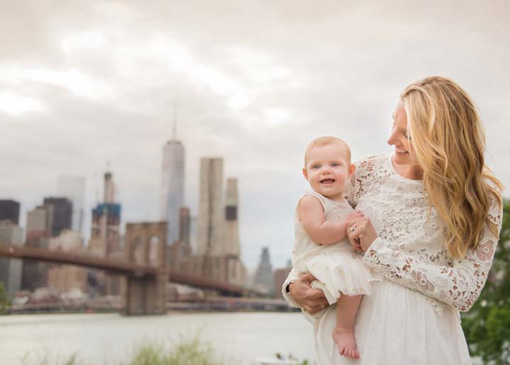 A woman with long blonde hair, wearing a white lace dress, stands outdoors, smiling while holding a baby dressed in a white outfit. The background features the Brooklyn Bridge and a cityscape with tall buildings under a cloudy sky.