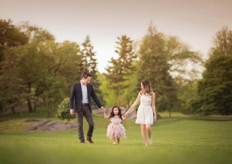 A family photographer captures a moment as a family of three, consisting of a man, woman, and young girl, walk hand in hand through a lush green park. The girl wears a pink dress while the adults wear formal attire. Trees and grass surround them under a soft, pastel sky.