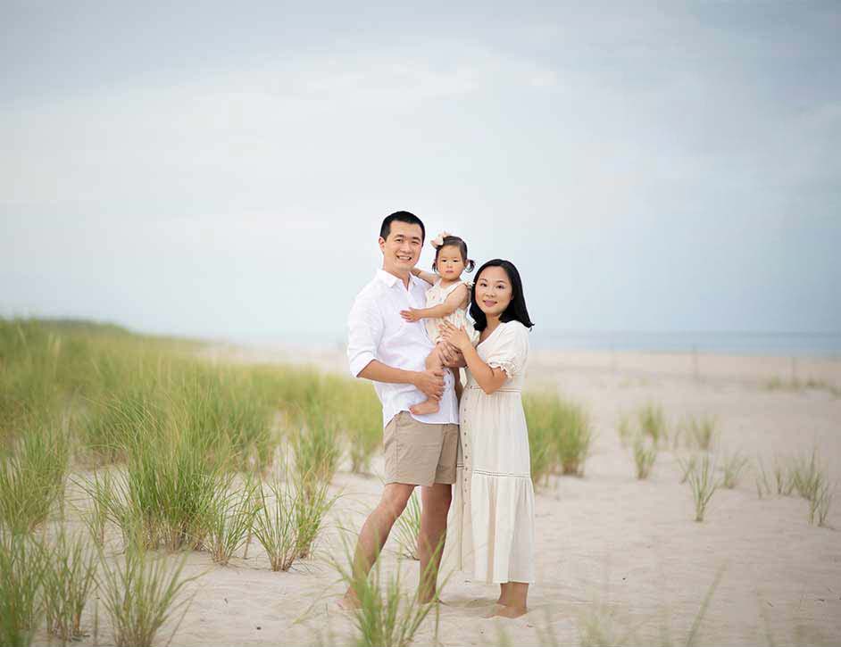 A family of three stands on a sandy NY beach with sparse grass. The father, in a white shirt and beige shorts, holds the toddler, who wears a white dress. The mother, in a light-colored dress, stands beside them, all smiling with the serene Southampton coastline in the background.