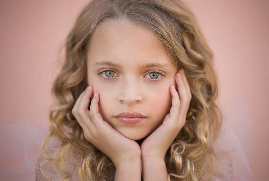 A young girl with long, curly blonde hair rests her chin on her hands, a thoughtful expression on her face. The background is a soft, peach color, capturing the innocence reminiscent of a baby photographer's enchanting work.