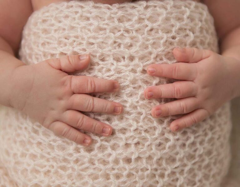 Close-up of an infant's hands gently resting on a soft, cream-colored knit blanket. The baby's fingers are touching the textured fabric, and the knitting pattern is clearly visible.