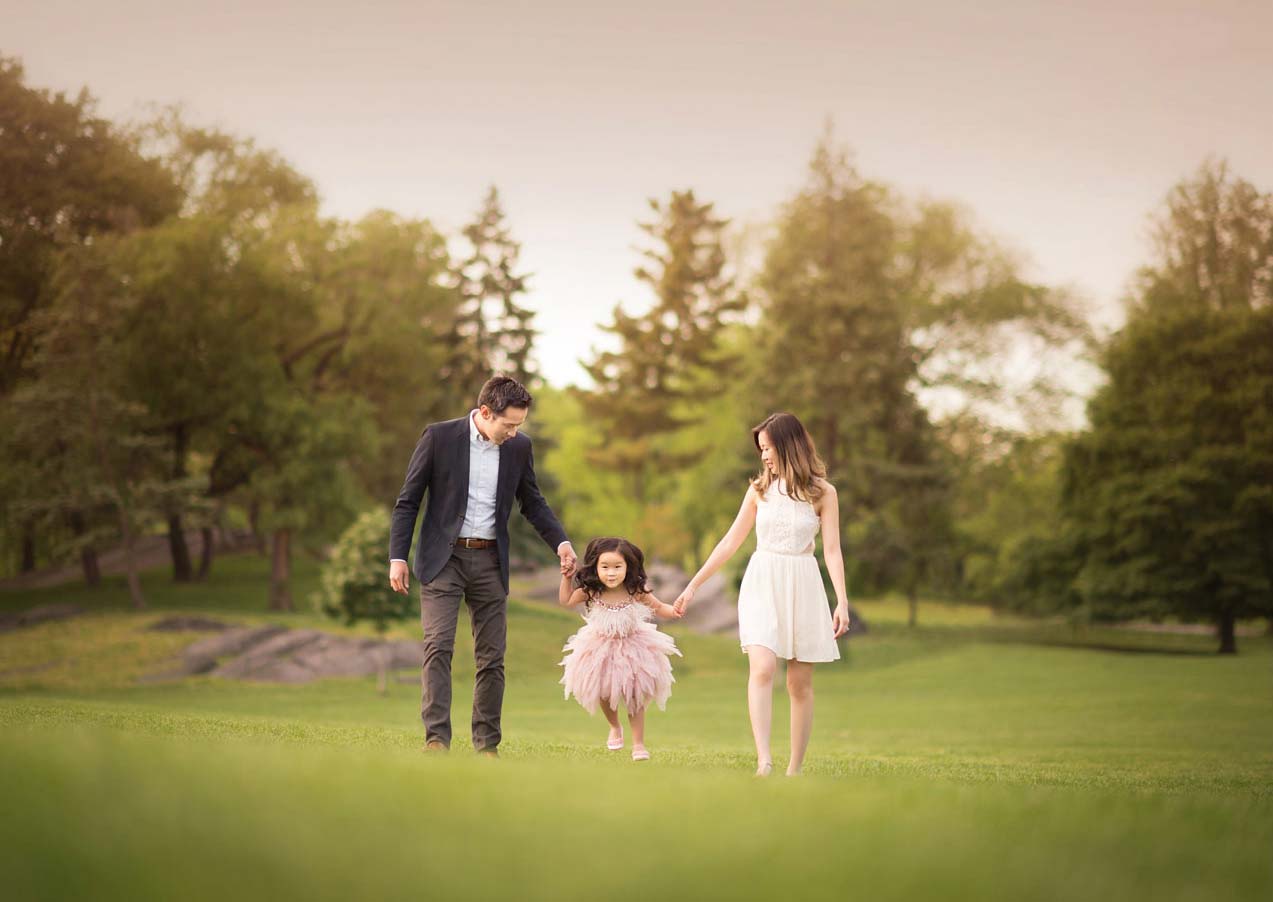 A married couple walks hand in hand with their young daughter in Central Park. The child wears a fluffy pink dress, and the parents are dressed elegantly. Trees and greenery surround them under a delicate, pastel sky. The family looks happy and joyful, enjoying their outdoor time.