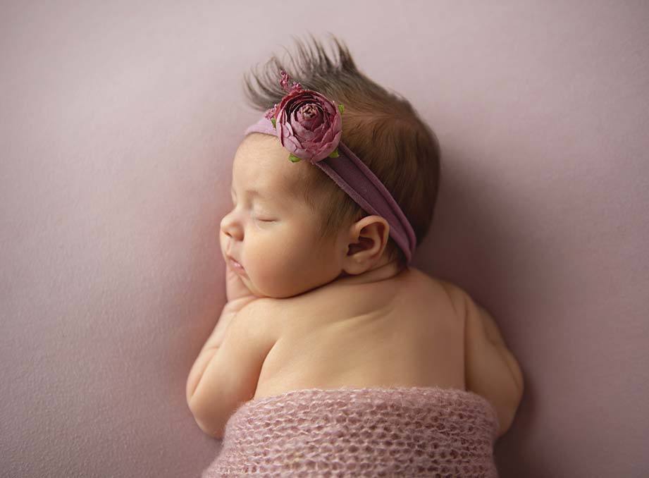 A sleeping baby with a small tuft of hair lies on their stomach on a soft, pastel pink blanket. The newborn wears a purple headband with a pink flower and is partially covered with a knitted pink wrap. The background, also pastel pink, creates the perfect scene for any newborn photographer.