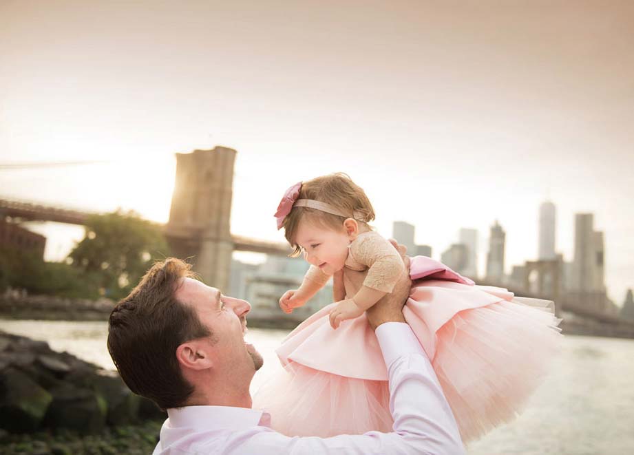 A father is joyfully lifting his baby daughter in the air near a waterfront with the NYC cityscape in the background, including a prominent bridge. The baby is wearing a pink dress with a bow in her hair, and both appear to be smiling and happy. The sky is overcast.