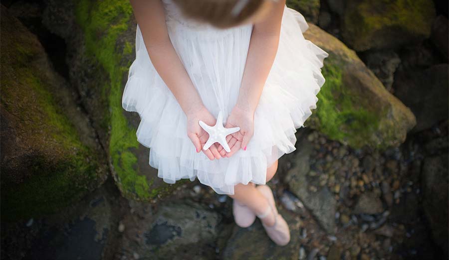 A girl in a white dress holds a starfish while sitting on moss-covered rocks. The perspective shows the girl from above, highlighting the starfish and the tulle fabric of the dress.