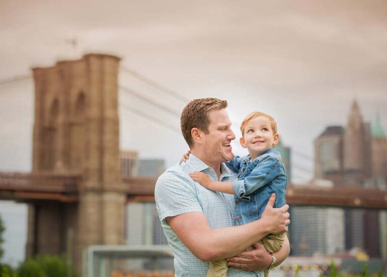 A man holds a young child in his arms while smiling. The child looks happy and is dressed in a denim jacket. They are outdoors, with a view of the Brooklyn Bridge and the New York City skyline in the background. The sky is overcast.
