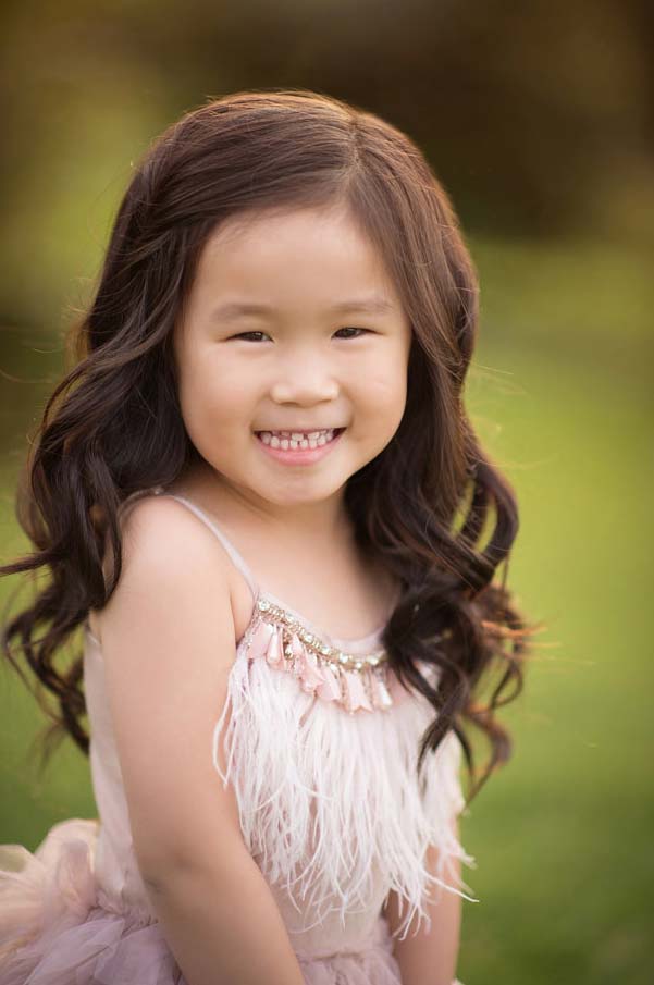 A young girl with long, wavy hair smiles brightly while posing outdoors. She is wearing a sleeveless dress adorned with feather-like embellishments and sparkly details, standing against a blurred background of greenery.