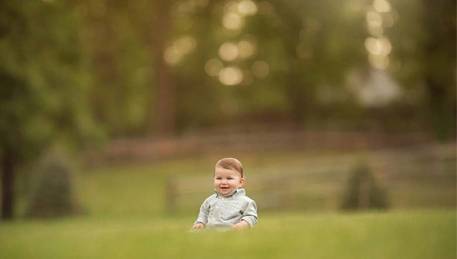 Smiling baby with short brown hair sits on lush green grass on an NJ farm, with a blurred background of trees and a fence under warm natural light. The baby wears a grey shirt, and the scene exudes a peaceful, serene atmosphere.