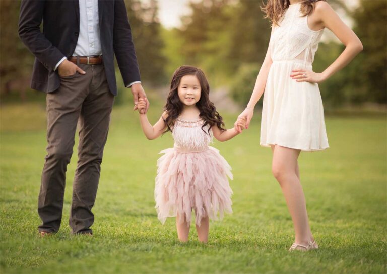 A young girl in a pink, feathered dress smiles, holding hands with two adults, who stand out of frame from the waist up. The group is standing on grass with trees and a blurred background. The adults are wearing casual yet stylish clothing.