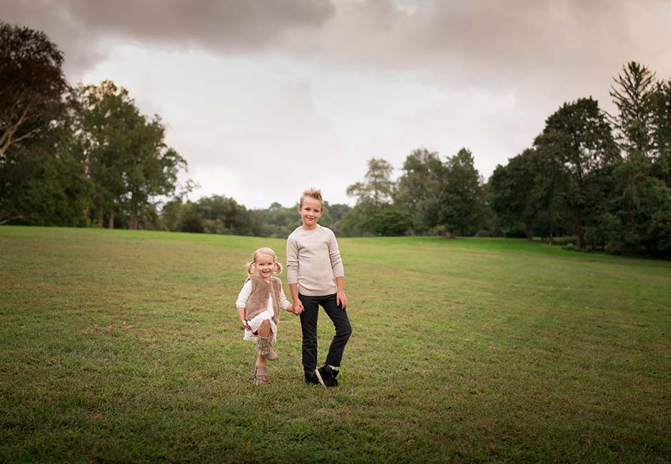 Two siblings are standing on a grassy field with trees in the background under a cloudy sky. The younger child on the left is playfully lifting one leg. Both are dressed in casual white tops and dark pants, smiling and looking at the camera.