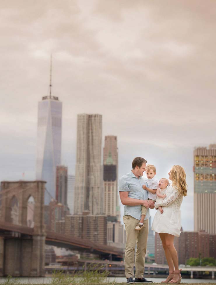 A family of four stands near the Brooklyn Bridge with the Manhattan skyline in the background, capturing a perfect moment of family photography. The parents, dressed in light summer clothing, hold their two young children and smile at each other under a cloudy sky.