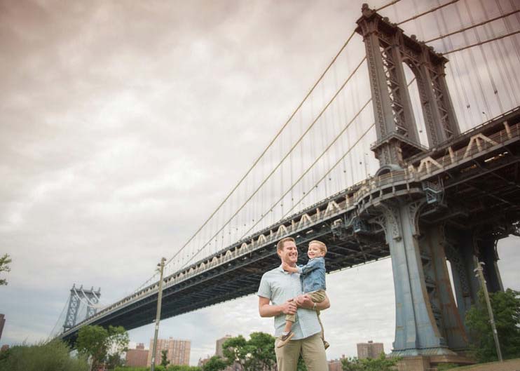 A man holding a young child stands in front of Manhattan Bridge, smiling. The sky is overcast, and there are trees and buildings visible in the background.