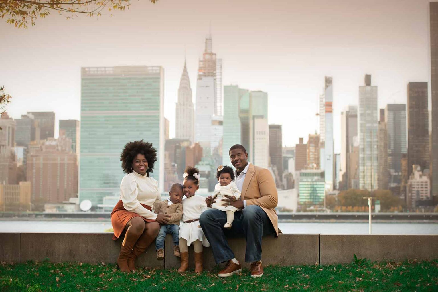 A family of five poses outdoors in front of the NYC skyline. The parents sit on a low wall, holding two young children, while a third child stands between them. They are all dressed in coordinated neutral-toned outfits, perfectly captured by a family photographer with the city's skyscrapers visible in the background.
