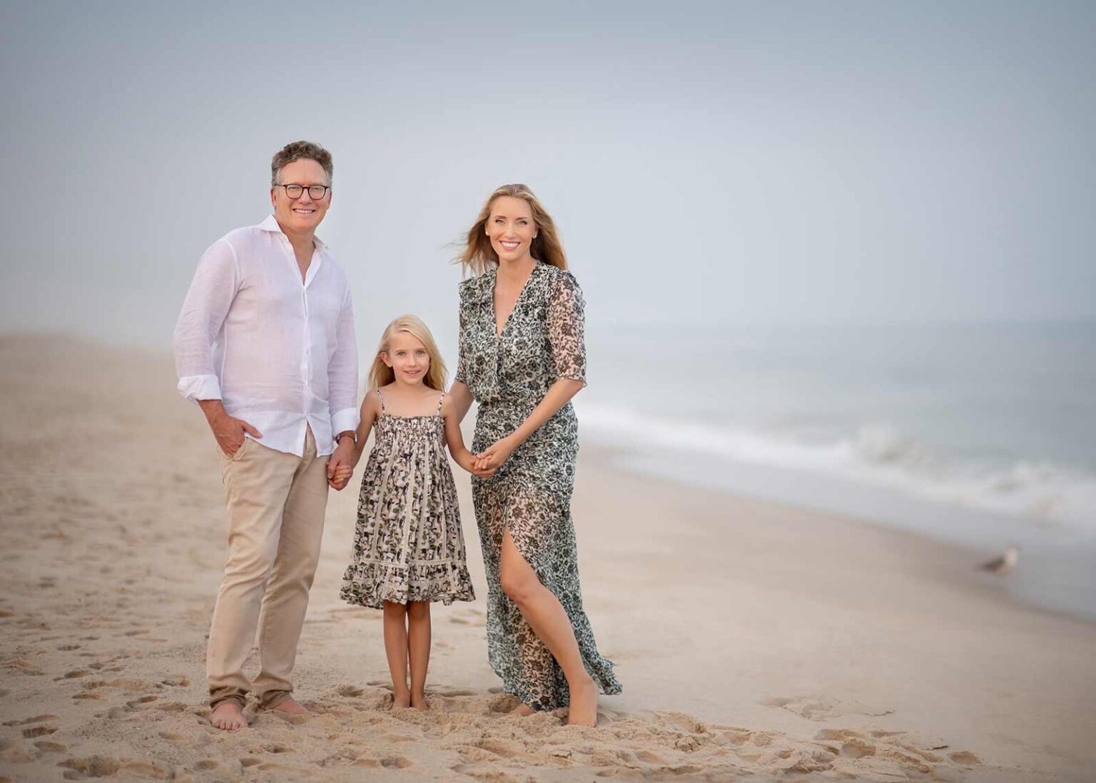 A family of three stands smiling on a sandy Coney Island Beach near the water. The man wears a white shirt and beige pants, the woman wears a flowing patterned dress, and the young girl wears a floral dress. The sky is clear, and a seagull is visible in the background.