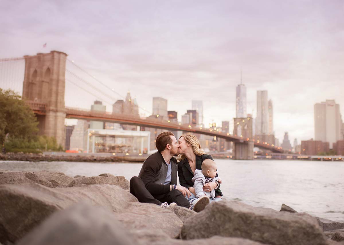 A family sits on rocks beside a river, in front of NYC cityscape dominated by the Brooklyn Bridge. They share a kiss while holding their baby. The sky is overcast, casting a soft light over the scene as the baby’s face turns slightly away, gazing at the water.