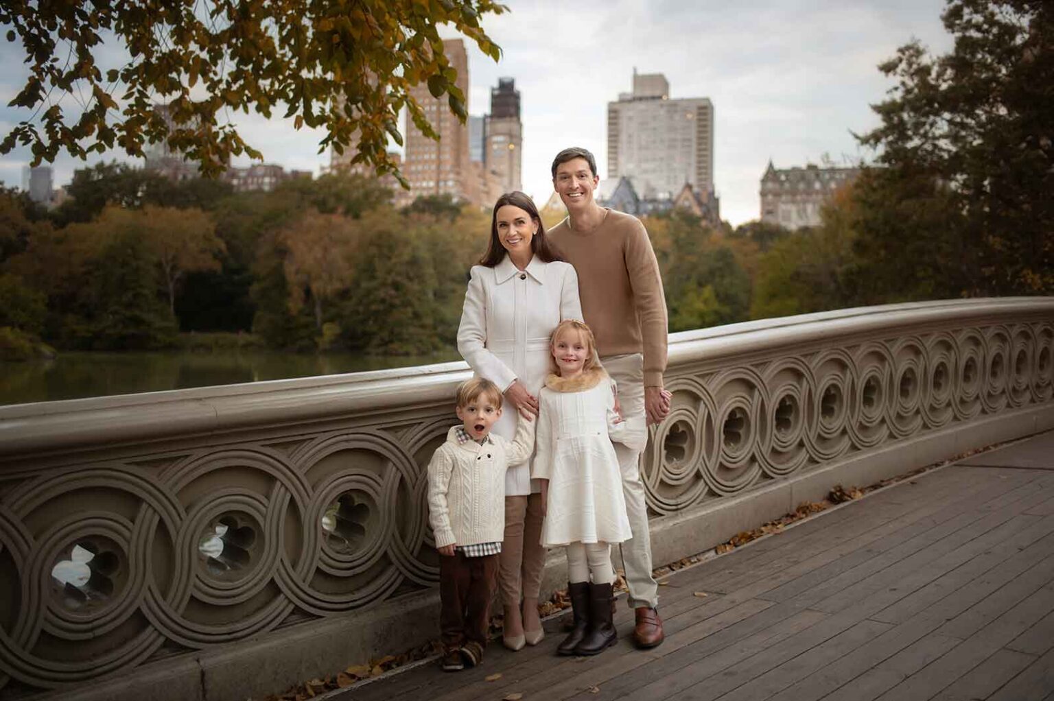 A family of four stands on a scenic bridge with intricate railings during a cherished family photoshoot in Central Park. The parents, dressed in autumn attire, smile at the camera while their two young children, a boy and a girl, stand in front. Trees and city buildings paint the background.