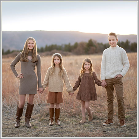 Four siblings stand holding hands in a field at sunset. They are dressed in autumnal clothing, with the boy on the far right wearing a white sweater and the three girls wearing brown and beige dresses and sweaters, reminiscent of cozy Hamptons attire.