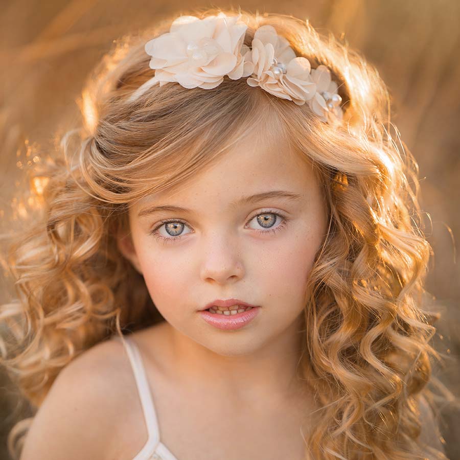 A young girl with long, curly blonde hair wears a white floral headpiece and a white dress. In this closeup shot by a family photographer, she looks directly at the camera with a soft expression. The background is blurred, featuring warm-toned, sunlit foliage. The image has a dreamy, golden-hour feel.