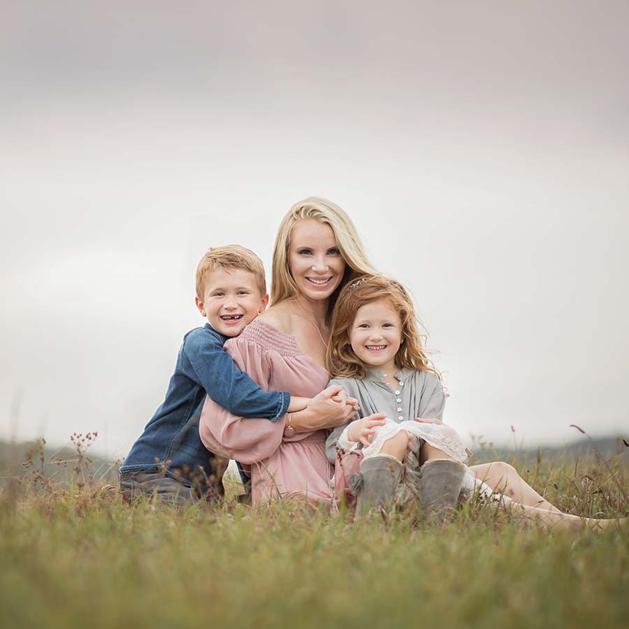 A mother, seated on grass, is smiling warmly with her young son and daughter during a family photoshoot. The boy on her left is wearing a blue shirt and laughing, while the girl on her right in a grey dress is smiling. The sky in the background is overcast, giving the scene a soft, serene ambiance.