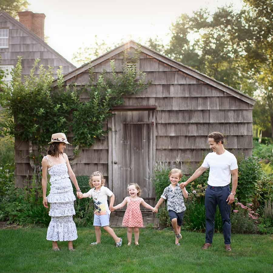 A family of five holds hands and stands on a grassy lawn in front of a rustic wooden shed, reminiscent of summer days in the Hamptons. The parents, both casually dressed, stand on either side of their children. The mother wears a white dress and hat. The children wear summer outfits and smile brightly.