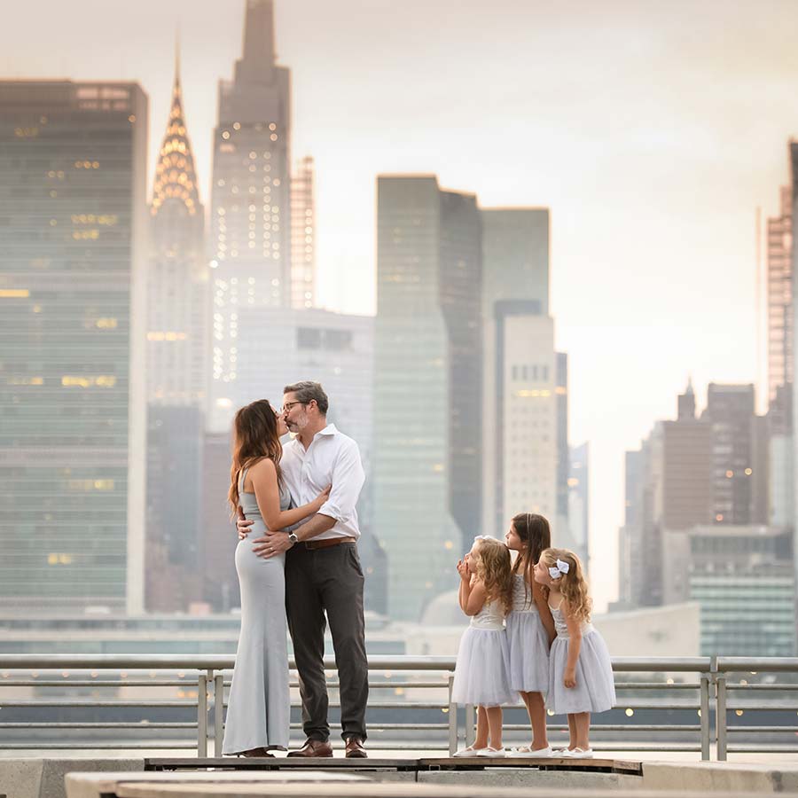 A couple stands on a rooftop with the NYC skyline in the background. They are embracing and gazing at each other affectionately. Nearby, three young girls in white dresses look up at the couple. This serene moment captures perfect family photography against a romantic urban backdrop.