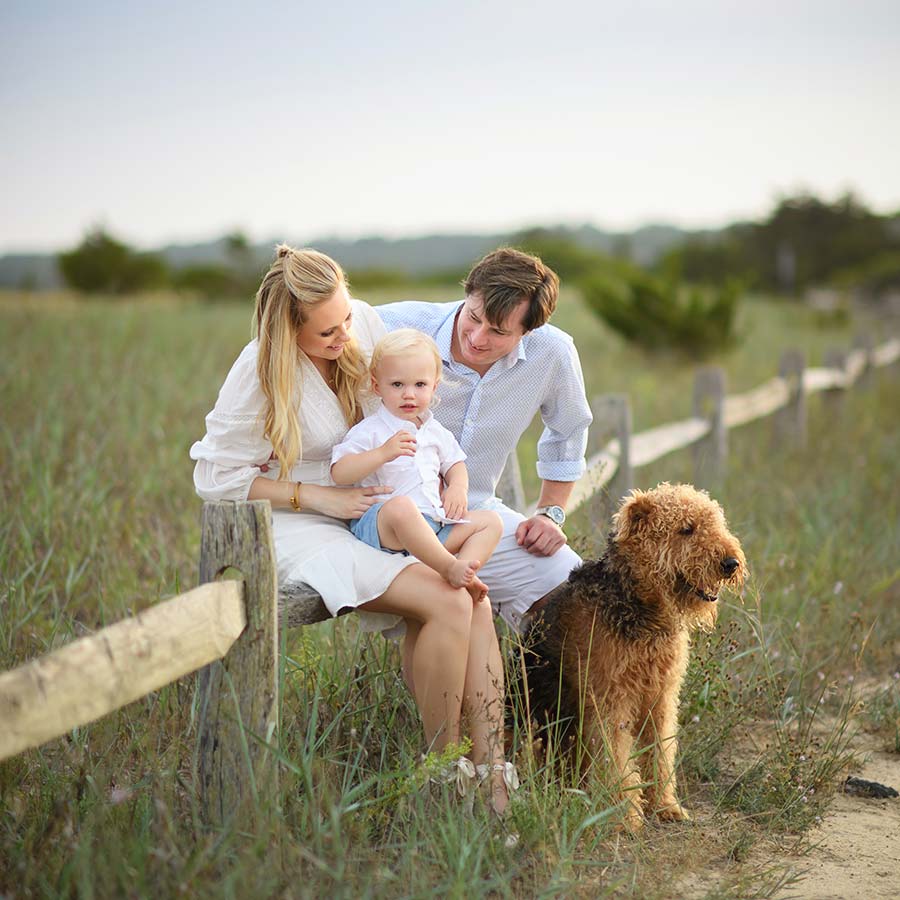 A family of three sits on a wooden fence in a grassy field near Southampton NY, captured beautifully by their family photographer. The young child, held by the parents, smiles brightly as their large, brown dog sits nearby. The parents' warm, engaging expressions create a loving and joyful atmosphere.