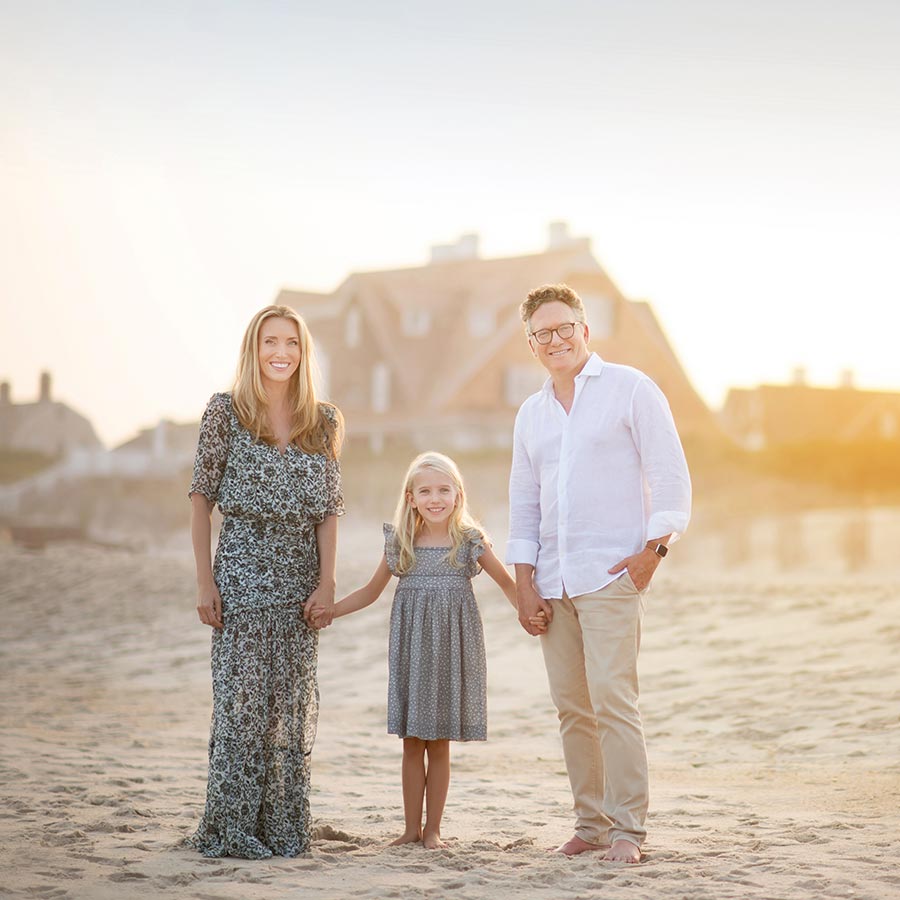A Hamptons family of three stands on a sandy shoreline at sunset, holding hands and smiling at the camera. The woman on the left wears a patterned dress, the man on the right wears a white shirt and beige pants, and the young girl in the middle wears a grey dress. Houses are in the background.