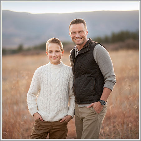 A father and his son stand side by side outdoors with mountains and a forest in the background. The man is wearing a grey sweater and black vest, while the boy is dressed in a white sweater. They are both smiling and have their hands in their pockets.