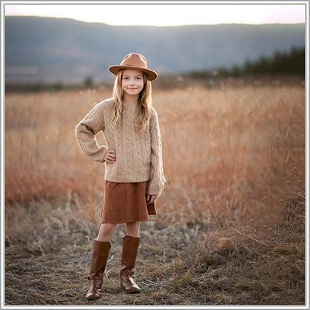 A young girl stands confidently in a field in the Hamptons. She is wearing a tan sweater, brown skirt, tan hat, and brown boots. Her hands are on her hips, and she is smiling at the camera. The field is dry with autumn grasses, and the sky is overcast.