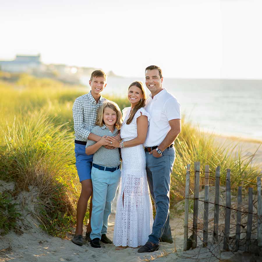 A family of four stands together on a sandy beach in Montauk NY, with tall grass on either side. The parents and two children are smiling at the camera. The sun is shining, and the ocean can be seen in the background, capturing a perfect moment of family photography.