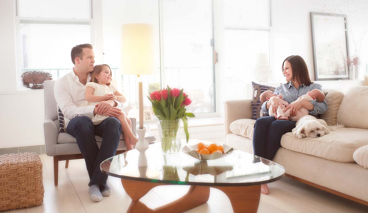 A NYC family is sitting in a bright living room. A man and a young girl are seated on a chair to the left, while a woman holding twin babies and a white dog are on a couch to the right. A round coffee table with flowers and a fruit bowl is in the center, creating a cozy inside home atmosphere.
