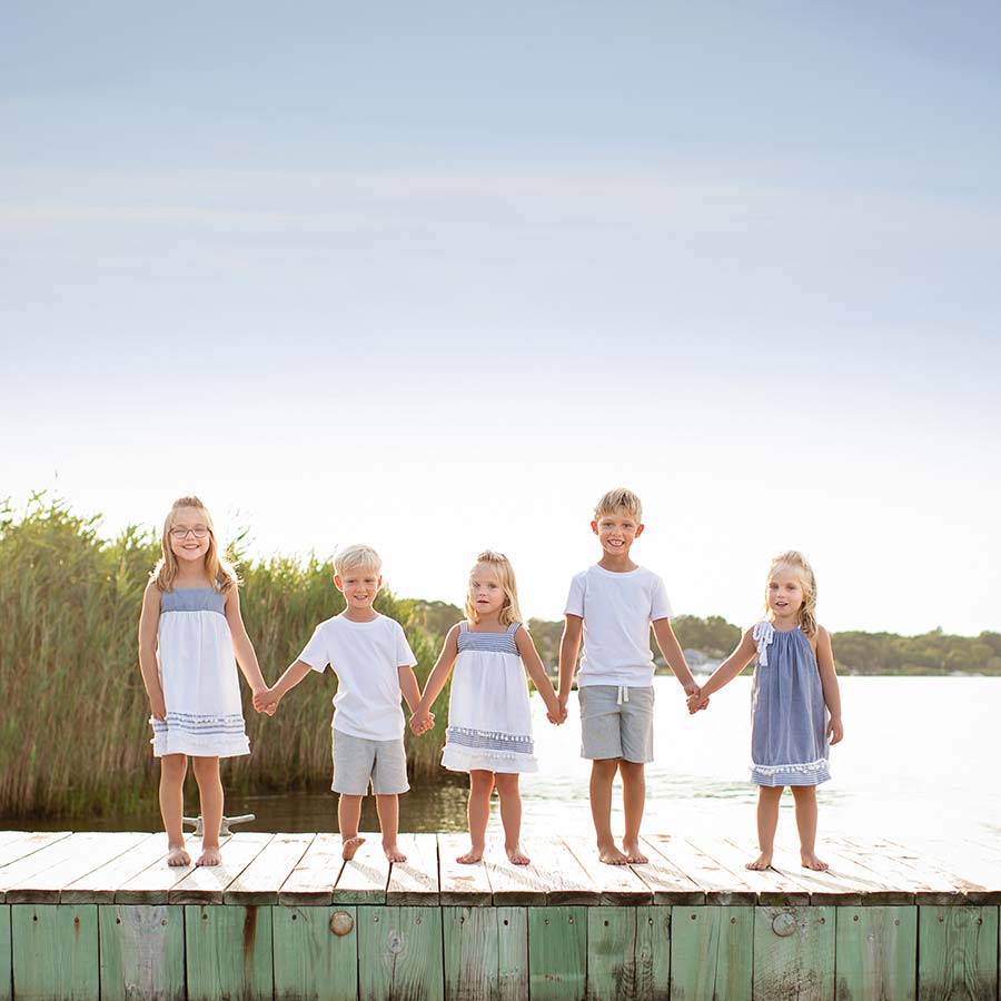 Five young children, three girls and two boys, hold hands while standing on a wooden dock in Sag Harbor, NY. They are dressed in summer clothes, and the background features reeds, water, and a clear sky.