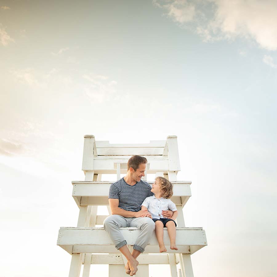 A father and son sit together on a large white lifeguard chair against a backdrop of a pale, partly cloudy sky at Easthampton Beach. The father is wearing a striped shirt and light pants, while the son is dressed in a white shirt and shorts. They are looking at each other and smiling.