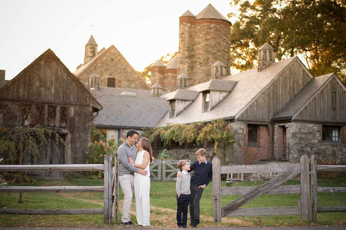 A family of four, two adults and two children, stands in front of a rustic stone farmhouse reminiscent of a Westchester county barn with a wooden fence in an idyllic countryside setting. The adults embrace while the two children stand nearby, with trees and a warm evening glow in the background.