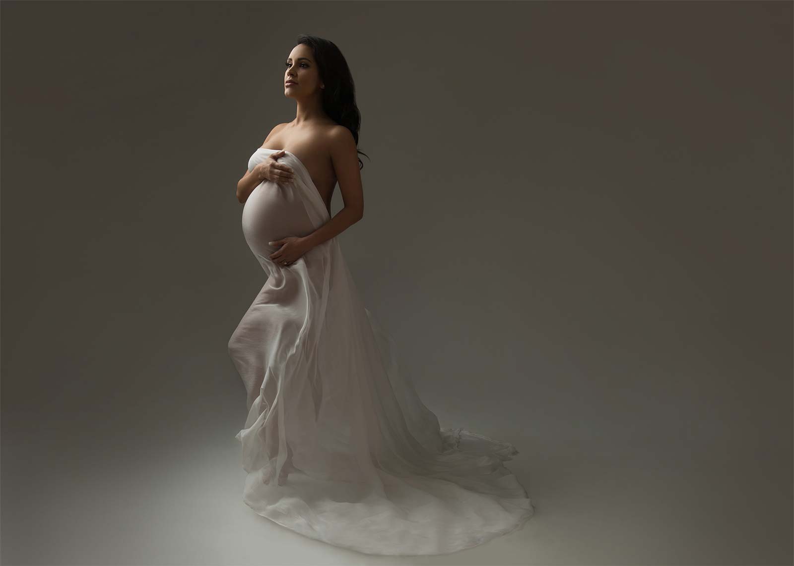A pregnant woman stands gracefully in a studio, draped in a flowy white fabric that cascades to the floor. She cradles her belly with both hands and looks serenely to the side, embodying the beauty of maternity. The background is soft and neutral, emphasizing the subject.