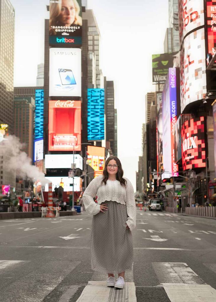 High school senior with brown hair and glasses stands in the middle of a Times Square NYC crosswalk, framed by bright digital billboards. She is wearing a white, long-sleeved top and a grey skirt. Perfect for a senior photoshoot backdrop, the iconic advertisements illuminate the scene beautifully.