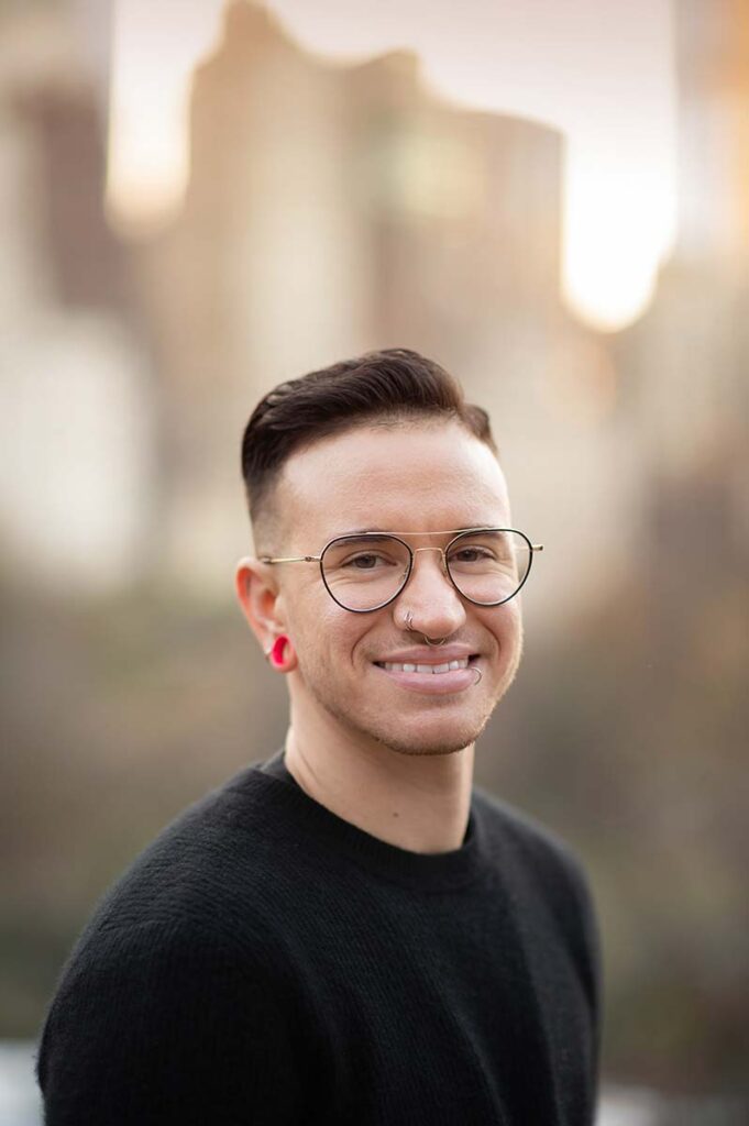 Young man with short dark hair, round glasses, a nose ring, and pink ear gauges smiles at the camera for his senior portrait. He is wearing a black top against a blurred outdoor background with buildings and trees.