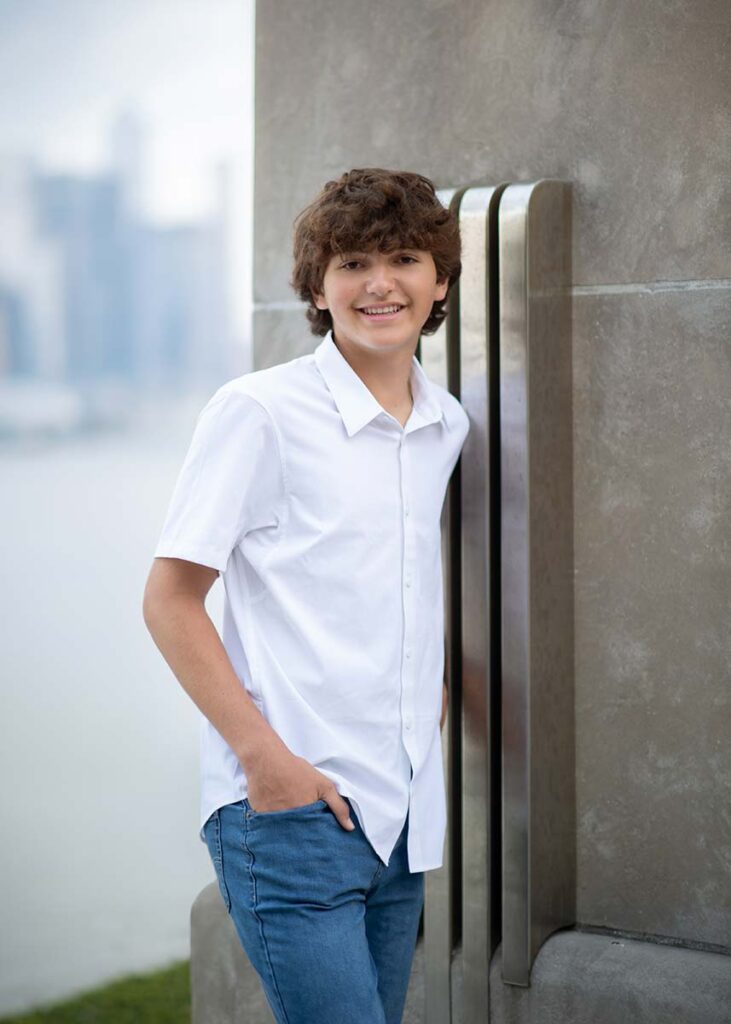 A young man with wavy hair is smiling and leaning against a structure, wearing a white shirt and blue jeans. The background is blurry, showcasing a cityscape with tall buildings and a body of water—a perfect setting for a high school senior photoshoot.