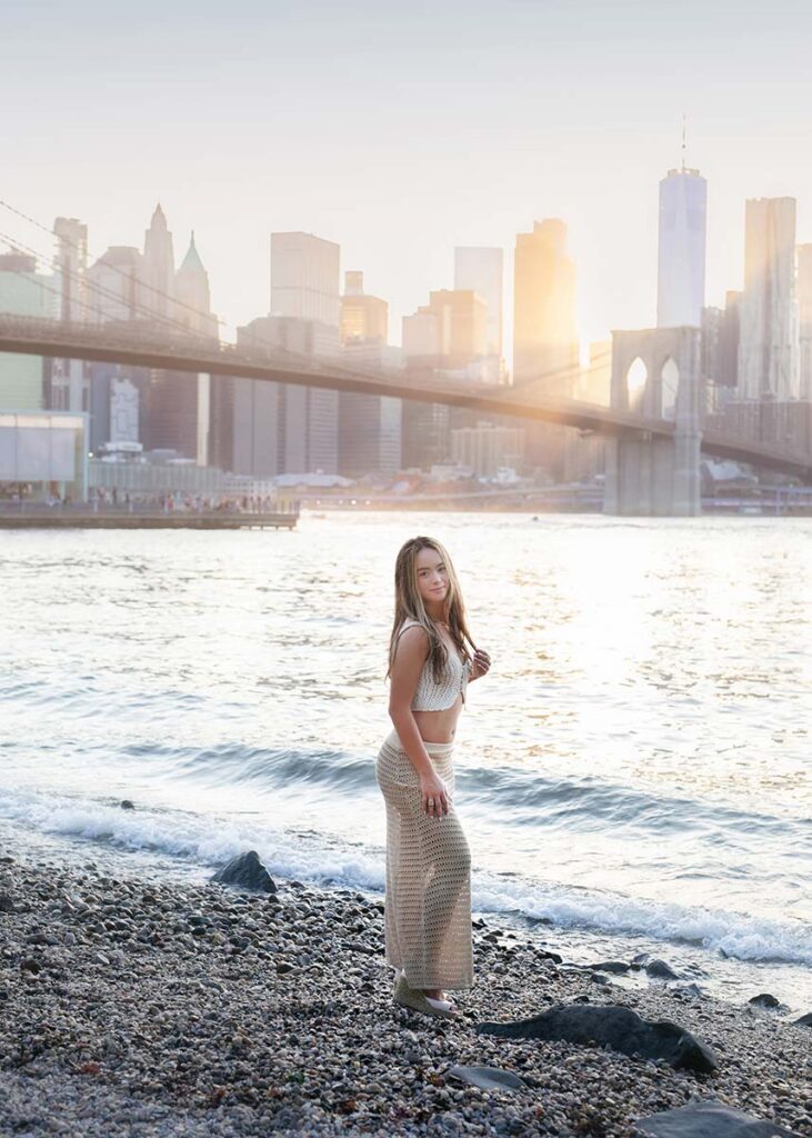 A young woman stands on a pebbled shoreline at sunset with the NYC waterfront and Brooklyn Bridge in the background. She is wearing a beige outfit and looking towards the camera as the warm sunlight bathes the scene, making her senior portrait serene and picturesque.